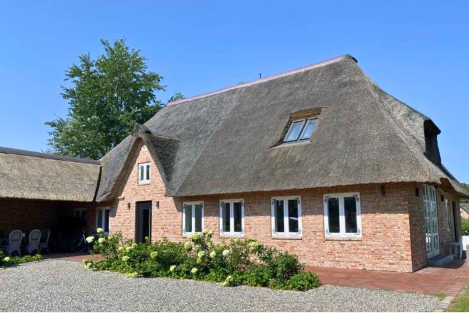 Traditionelles Reetdachhaus mit Garten und Fenstern, umgeben von Bäumen und Blumen, ideal für ländliche Architektur und Wohnprojekte.