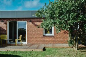 Moderne Backsteinwohnung mit großen Fenstern und einer Terrasse, umgeben von einem Baum und einem Tisch mit Stühlen.