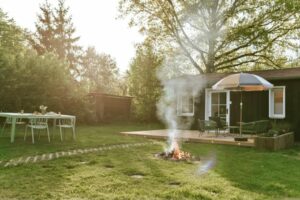 Ein Garten mit einer Feuerstelle, einem Sonnenschirm und Gartenmöbeln, umgeben von Bäumen und Gras.