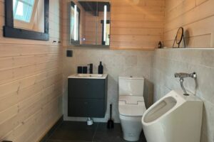 Ein modernes Badezimmer mit einem Waschbecken, WC und Urinal in einem Holzhaus, ausgestattet mit eleganten schwarzen Accessoires.