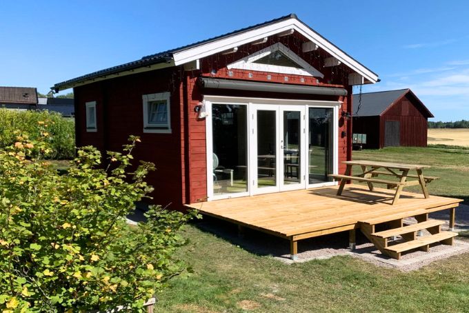 Rotes Gartenhaus mit Holzterrasse, großen Fenstern und Sitzgelegenheit im Freien, umgeben von Grünflächen.