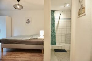 Schlafzimmer mit Bett, Nachttischlampe und angrenzendem Badezimmer mit Dusche und Fliesen. Ideal für moderne Wohnräume.